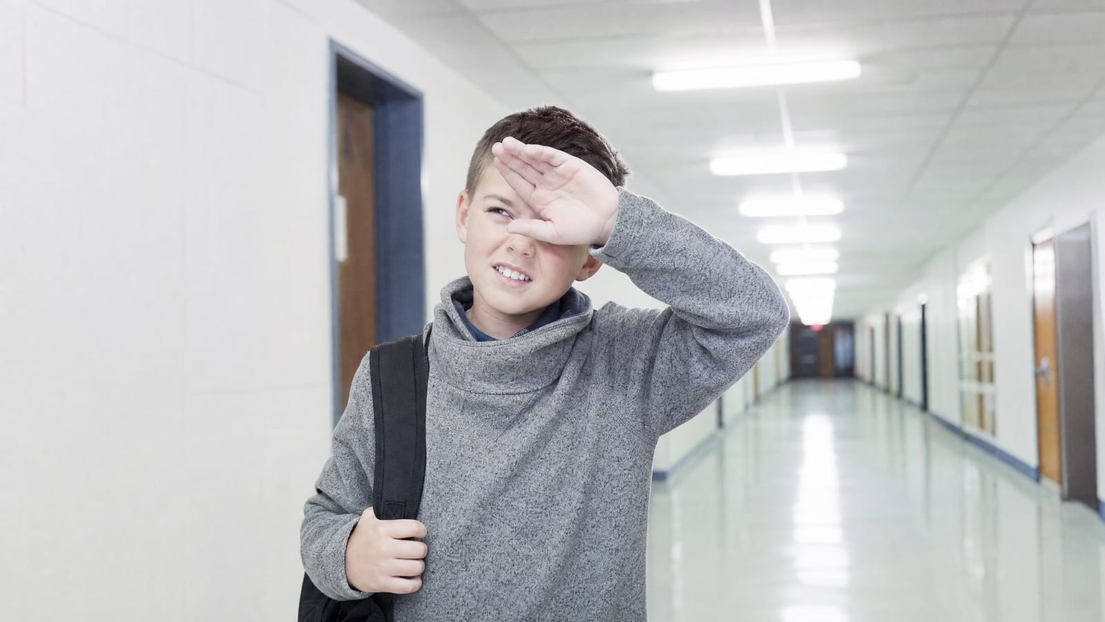 A child pausing in a bright school corridor, squinting and shielding their eyes with one raised hand against rows of harsh overhead fluorescent lighting reflecting off a polished floor