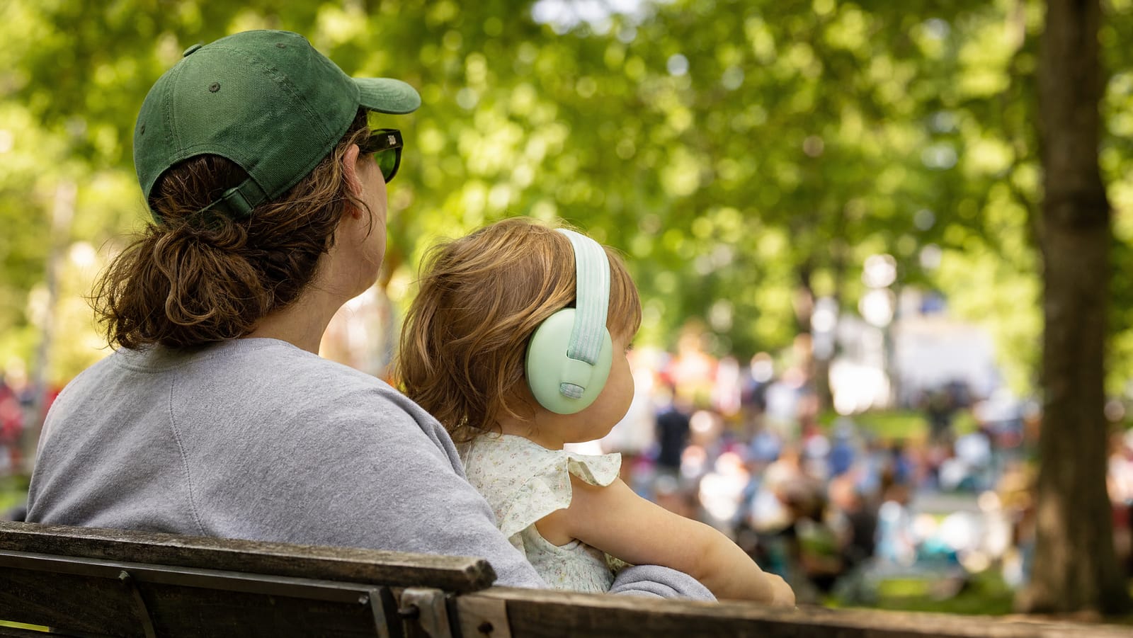 A toddler wearing soft mint padded ear defenders sitting calmly on a parent lap on a park bench, both facing away towards a softly blurred crowd at a sunlit outdoor event